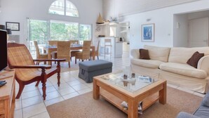 A spacious living room with a high ceiling, a dining area with wicker chairs, a sofa, and a glass coffee table.