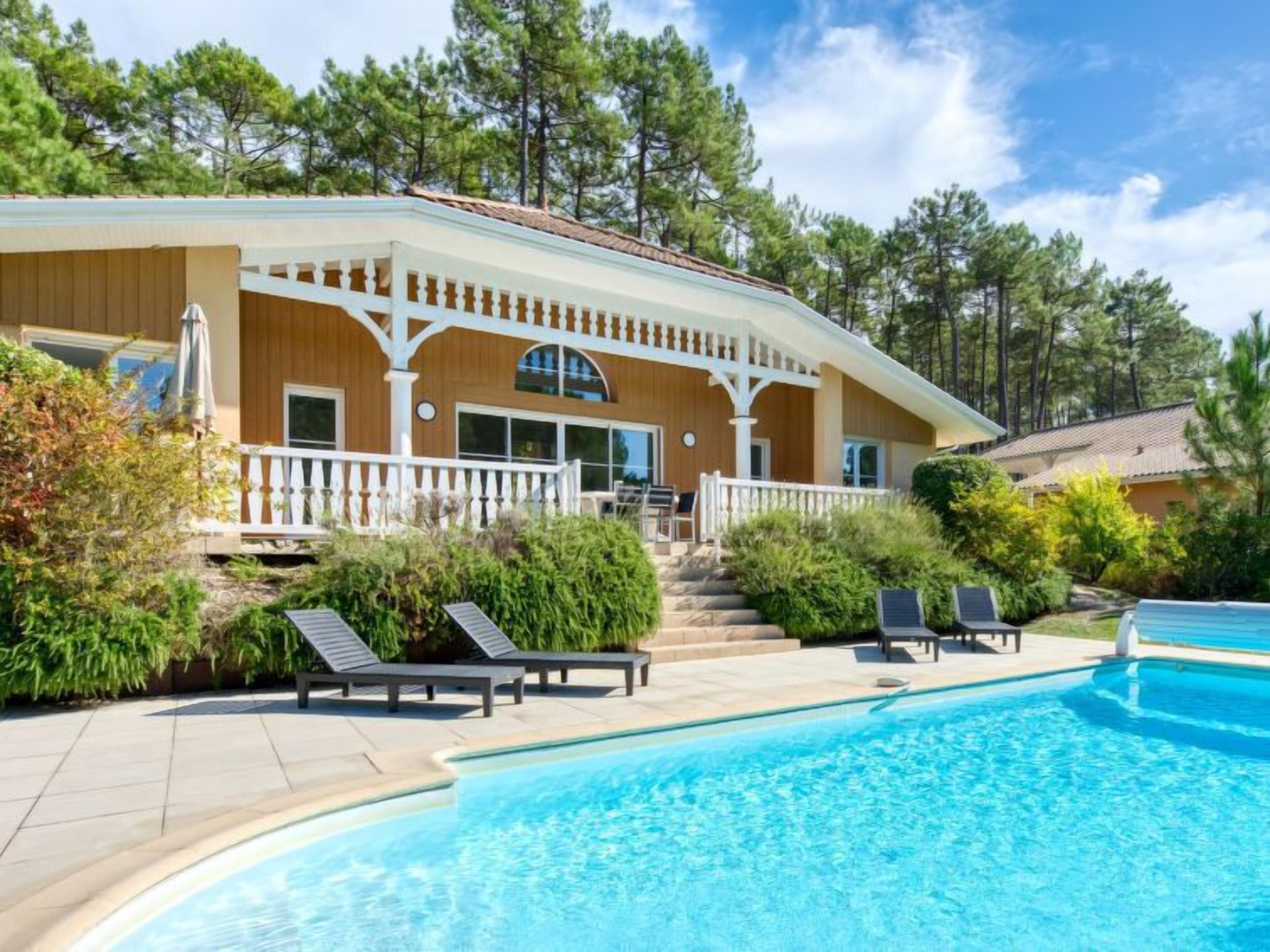 A two-story house with a pool, surrounded by trees and a patio area with lounge chairs.