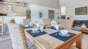 A modern living room with a dining area, featuring wicker furniture, a wooden dining table, and a view of the sea through large windows.