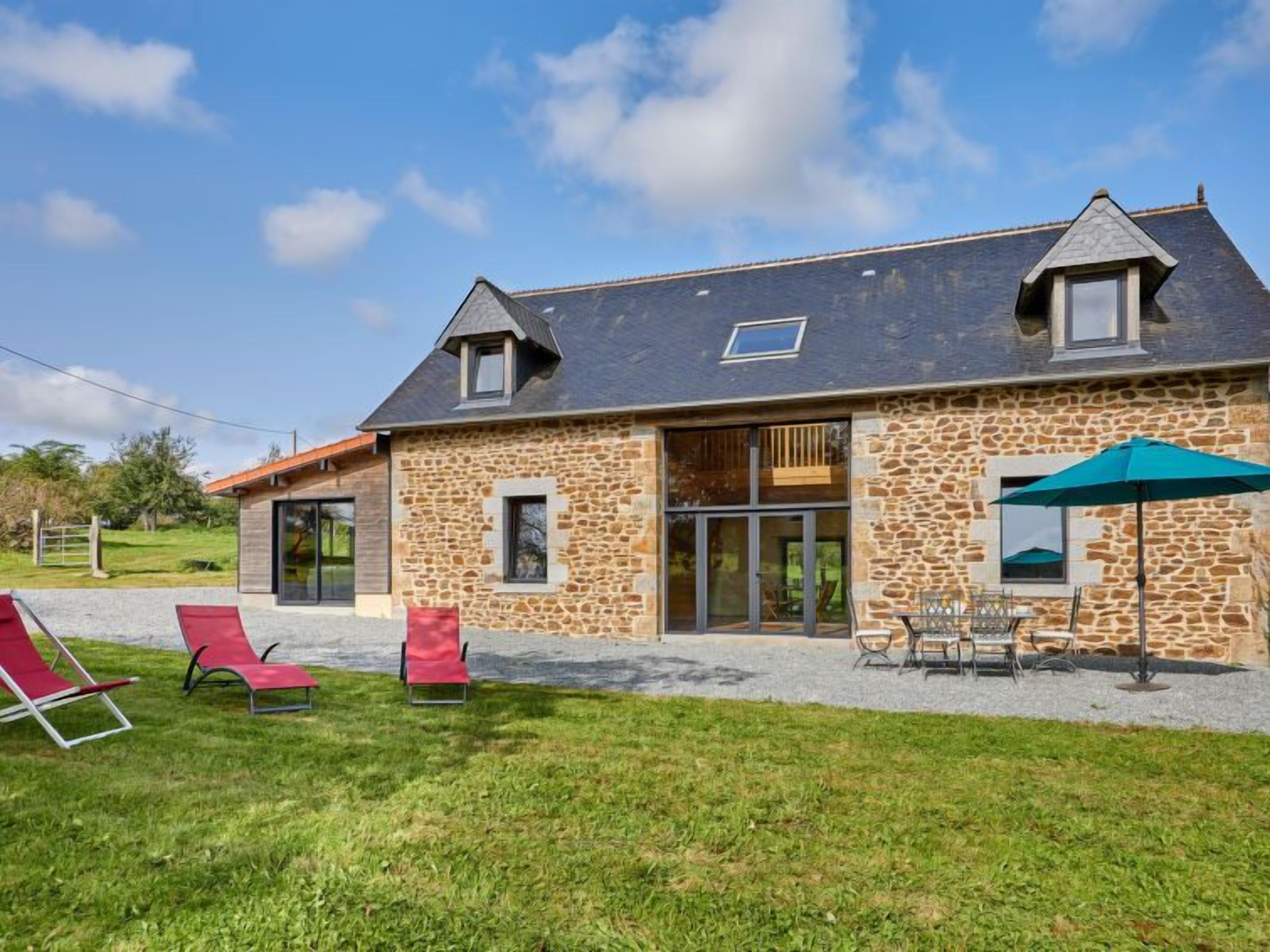A stone house with a large glass door, a covered patio with a table and chairs, and a grassy lawn with red lounge chairs.