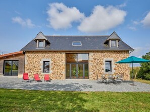 Une maison en pierre avec une grande porte d’entrée, deux fenêtres à l’étage et une terrasse aménagée avec du mobilier d’extérieur.