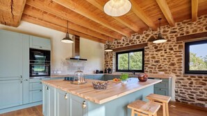 A kitchen with a central island, wooden ceiling beams, stone wall, and large windows. - La Favrais by Interhome (Saint aubin de terregatte)