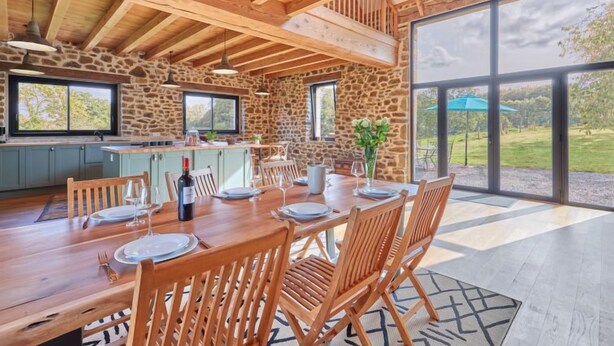 A spacious dining area with a wooden table set for four, stone walls, and large windows offering a view of the outdoors. - La Favrais by Interhome (Saint aubin de terregatte)