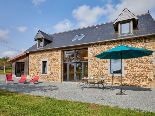 Une maison en pierre avec un toit en pente, un patio couvert avec des sièges d’extérieur et un grand parasol.