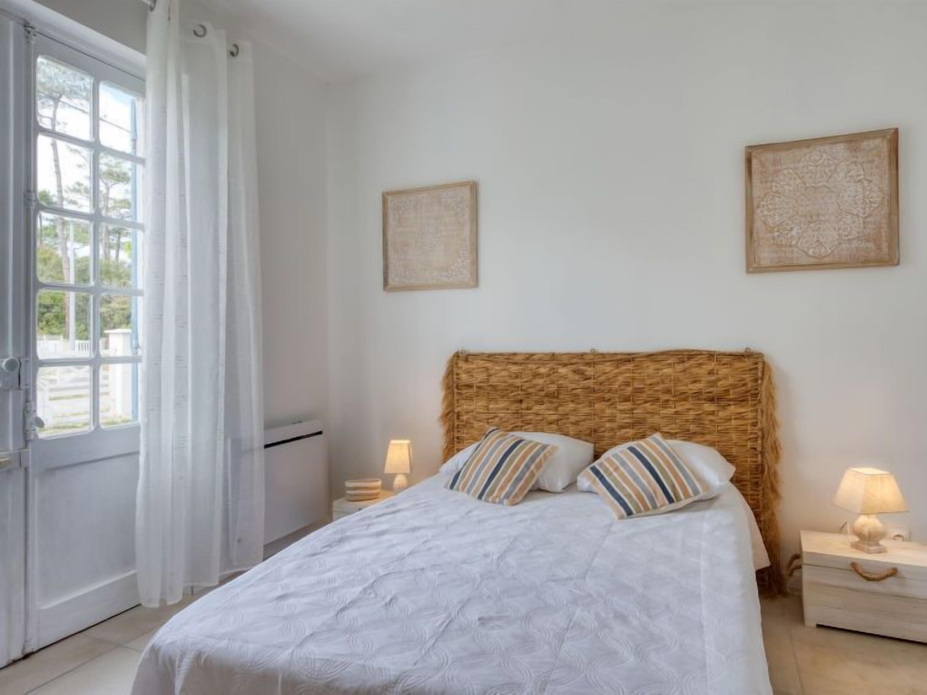 A bedroom with a large bed, two bedside tables, a window with curtains, and two wall-mounted artworks.