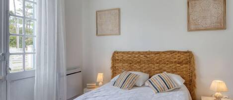 A bedroom with a large bed, two bedside tables, a window with curtains, and two wall-mounted artworks.