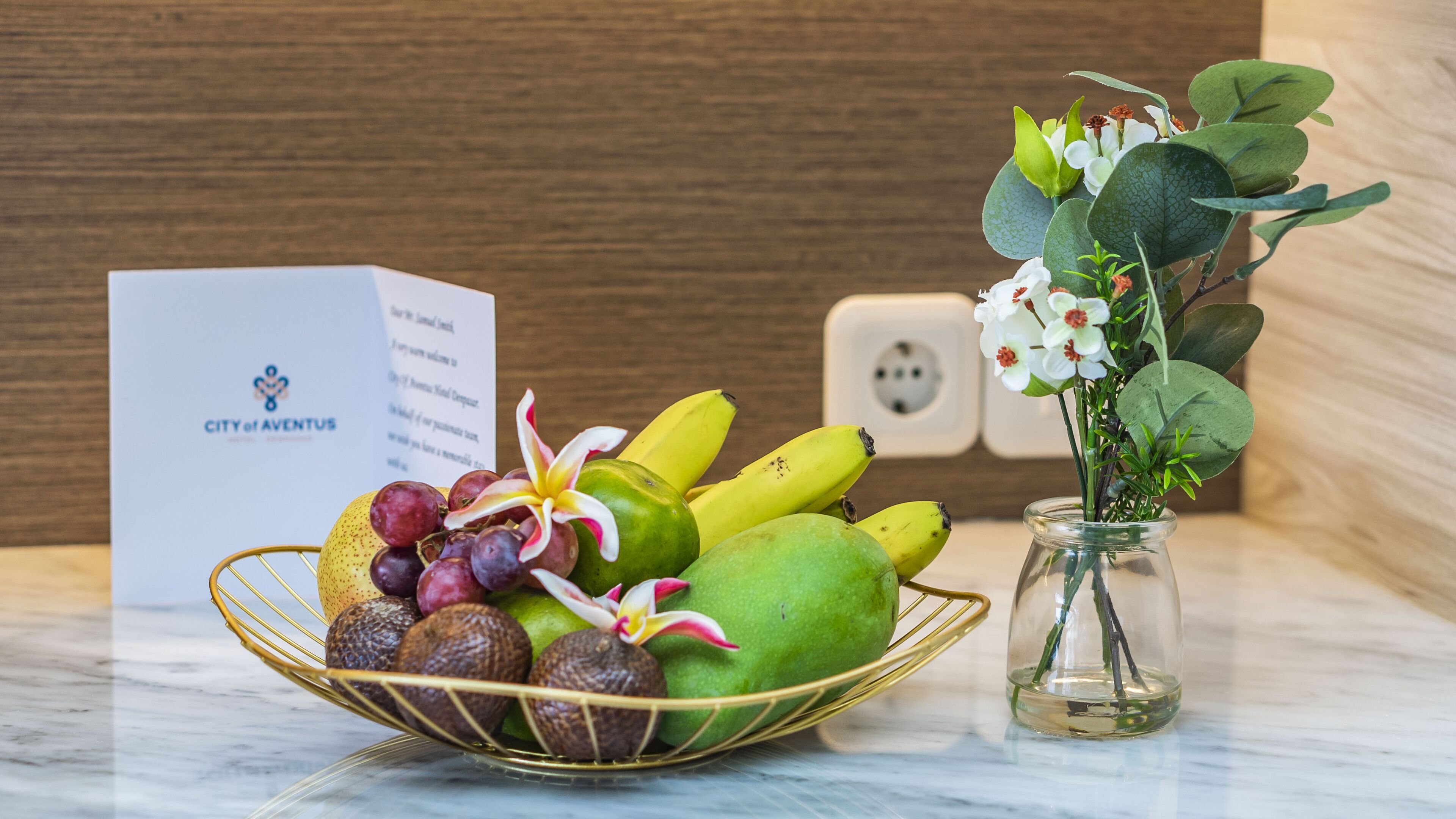 Una cesta de frutas con uvas, plátanos y una fruta verde, un jarrón con flores, y un cartel con el texto 