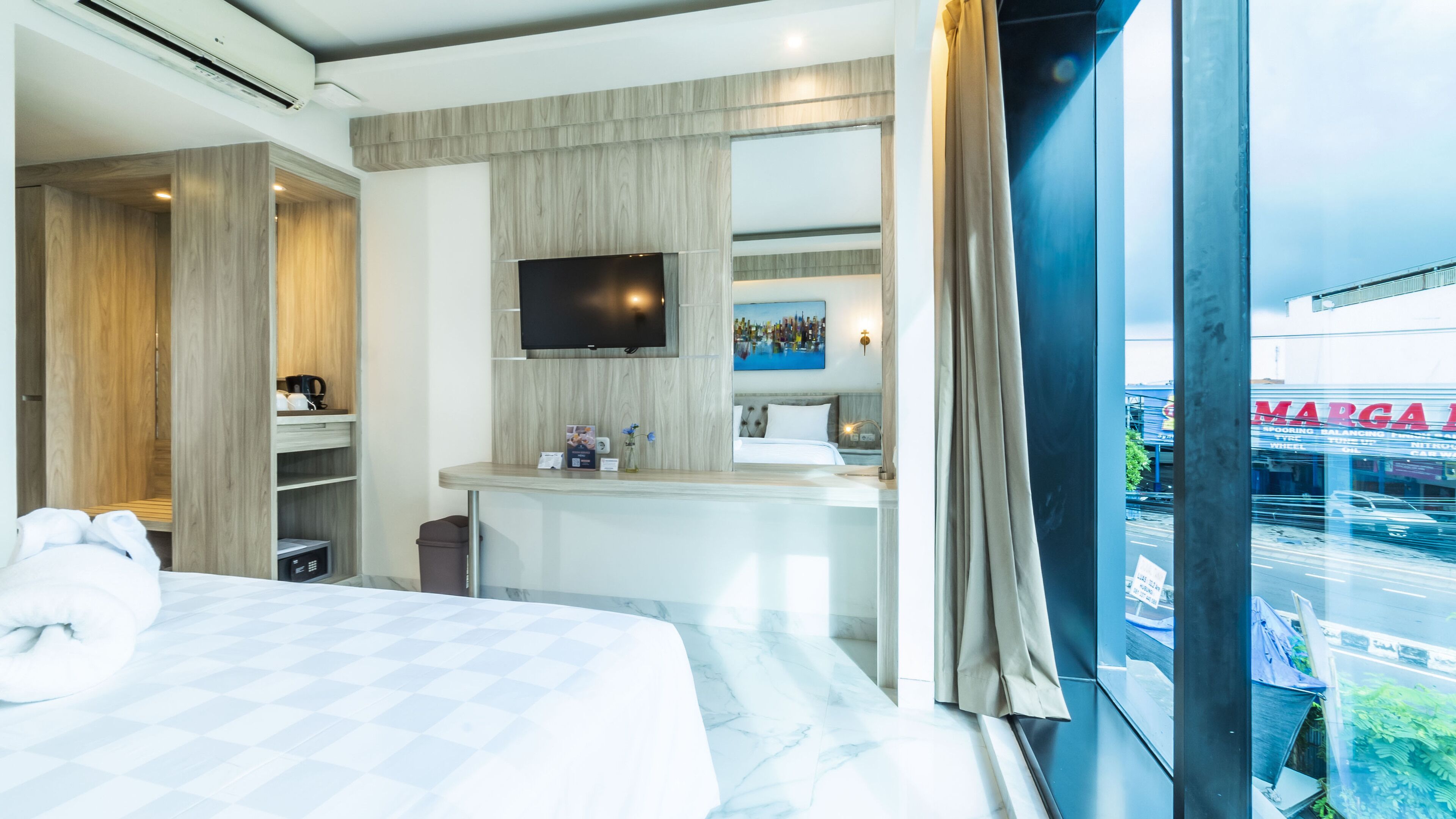 A hotel room with a bed, a TV mounted on the wall, a small desk with a chair, and a window with a view of a cityscape.