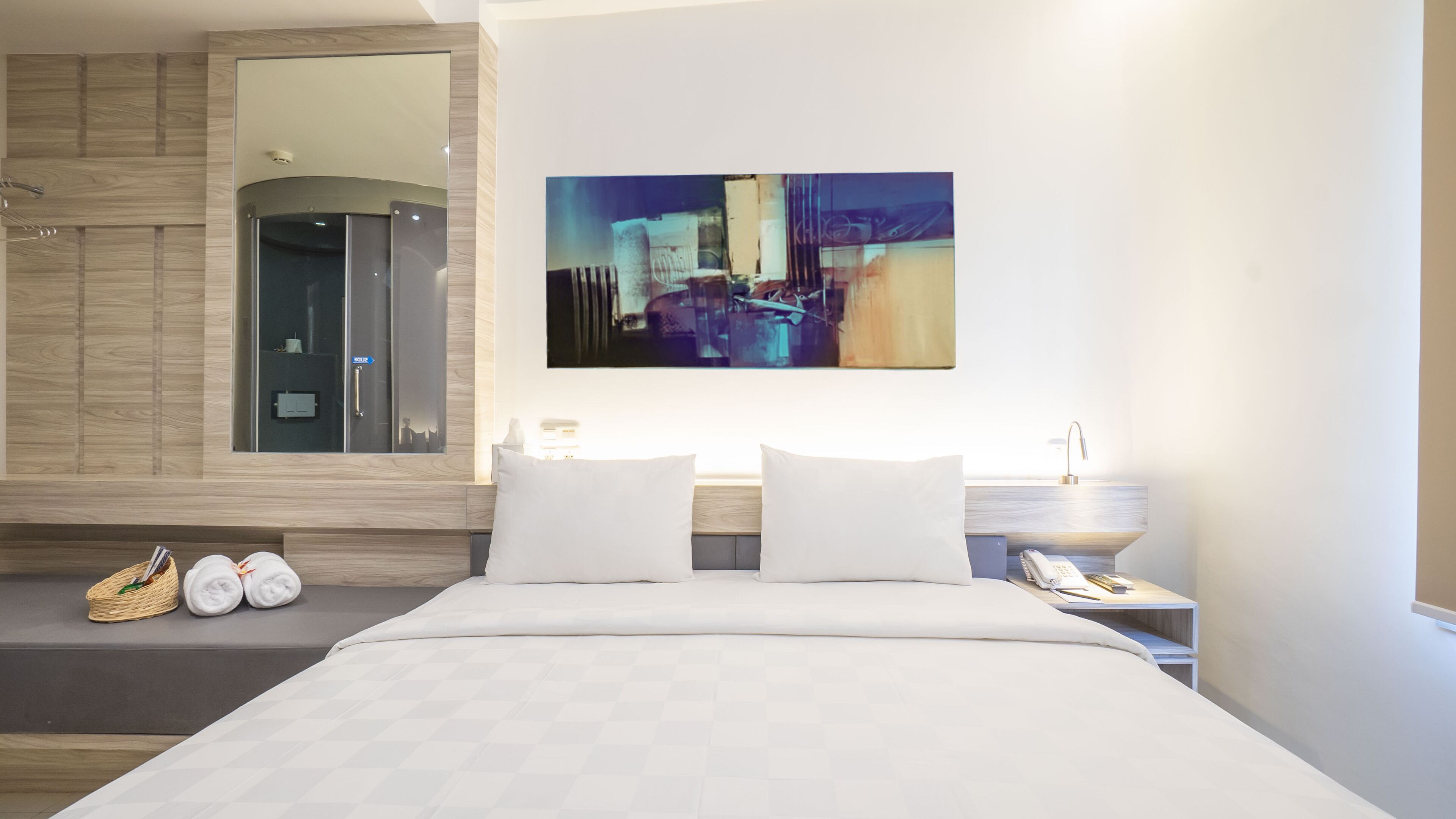 A modern hotel room with a large bed, a mounted artwork, a mirror, and a small side table with a lamp.