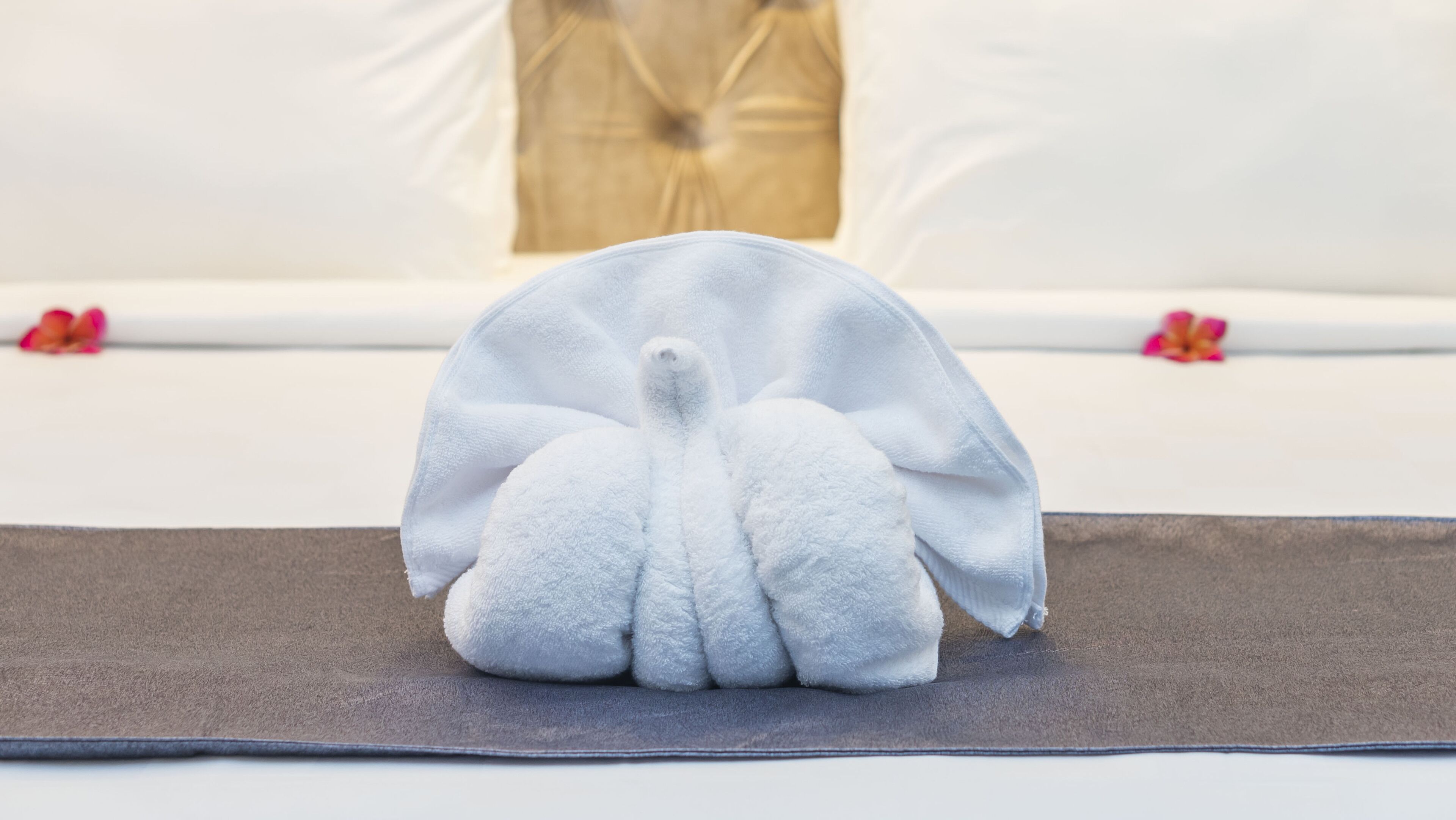 A white towel folded into a fan shape on a brown mat, with a white cushion and a beige pillow in the background.