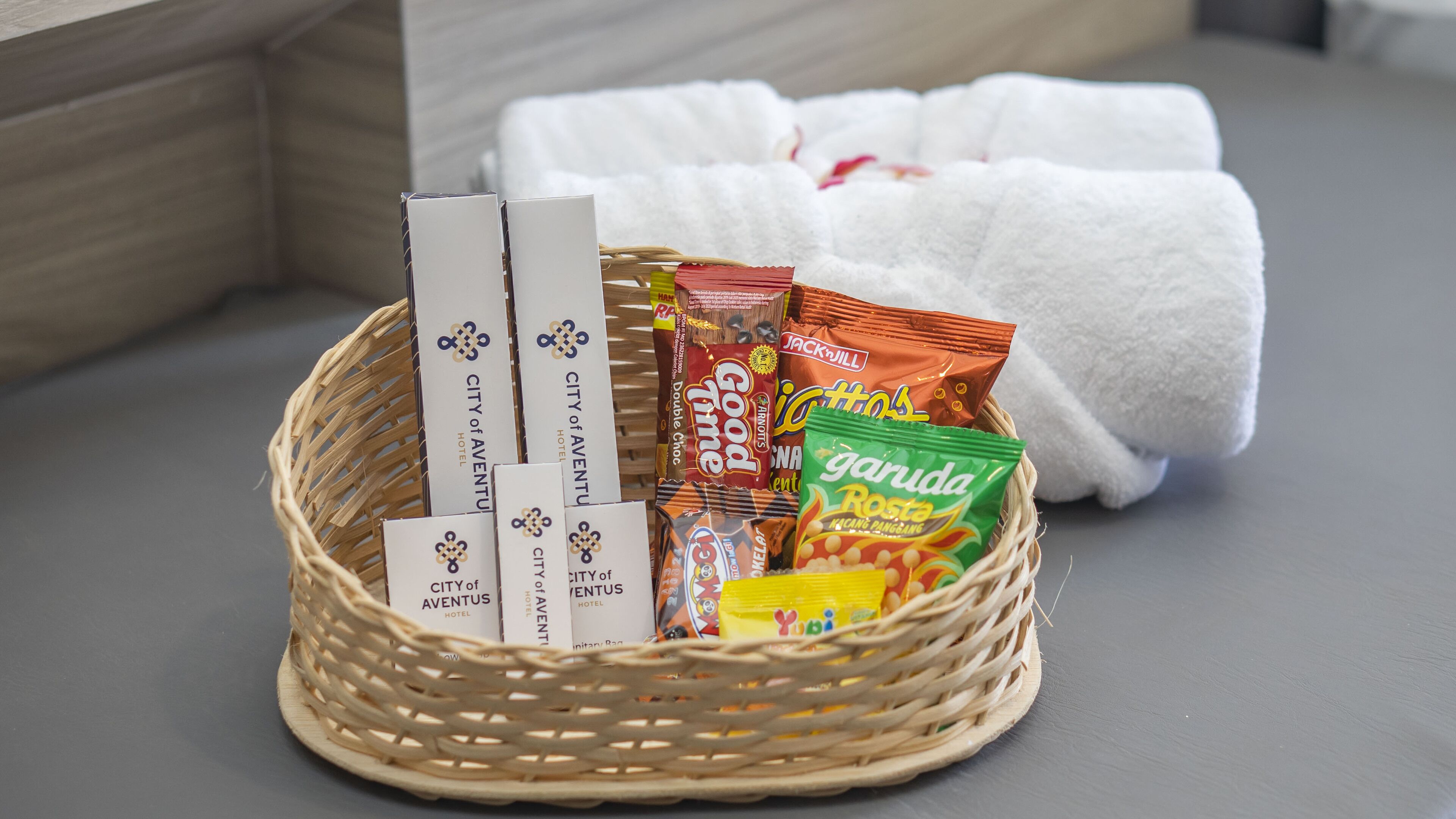 Una cesta de mimbre con folletos del hotel, aperitivos y una toalla blanca sobre una superficie gris.