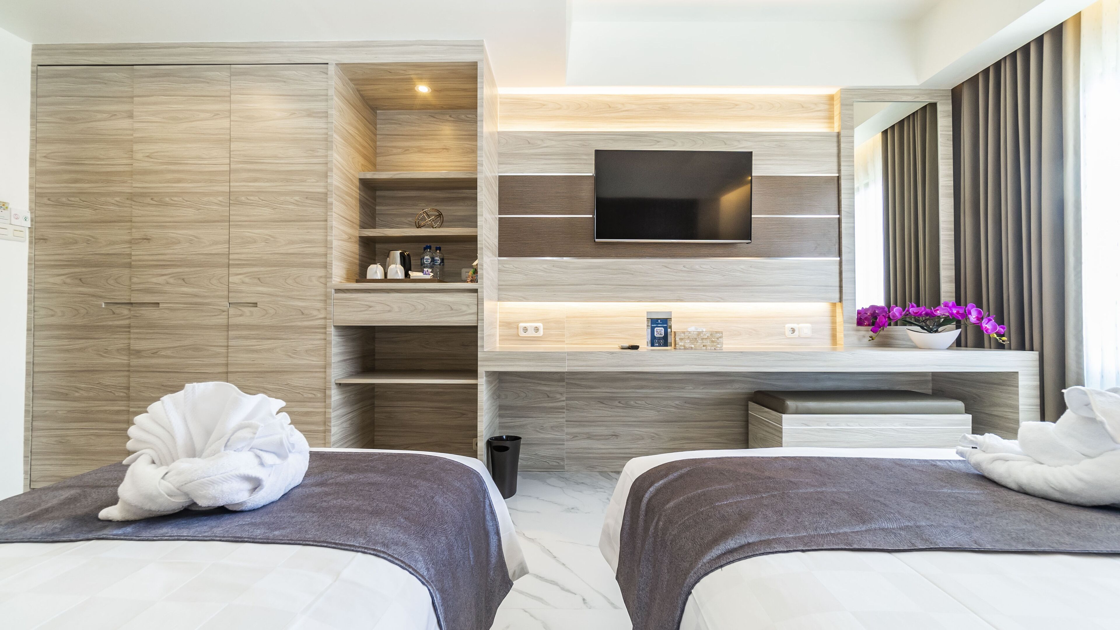 Een moderne hotelkamer met een houten wand, een flatscreen televisie, een klein plankje met toiletartikelen en twee bedden met handdoeken.