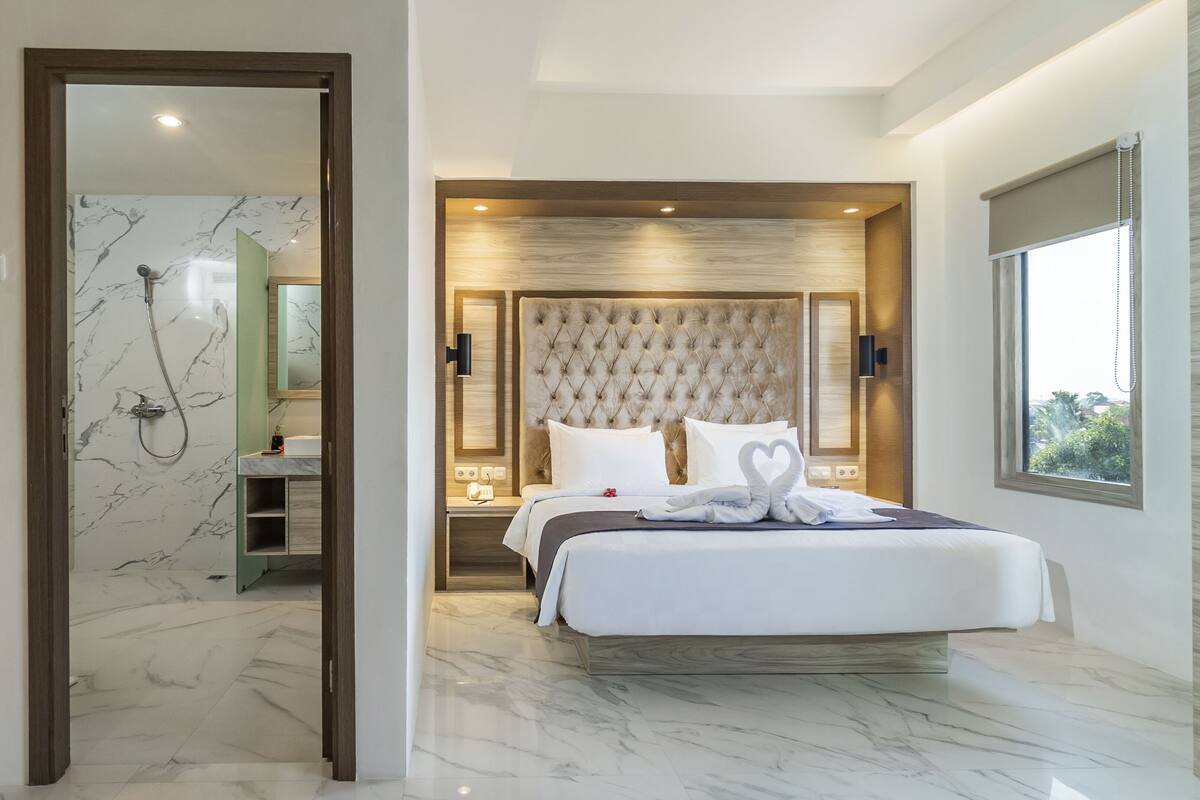 Ein modernes Hotelzimmer mit Marmorbad, einem großen Bett mit gepolstertem Kopfteil und Blick ins Freie durch ein Fenster.