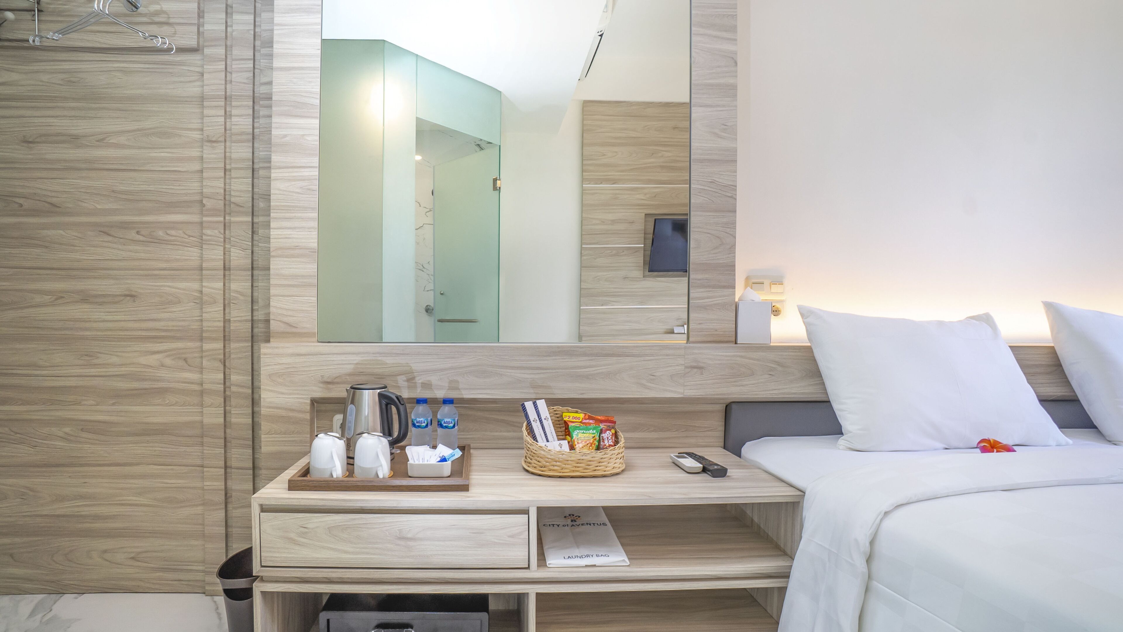 A hotel room with a wooden vanity, a mirror, a bed with white bedding, and a bedside table with toiletries and a remote control.