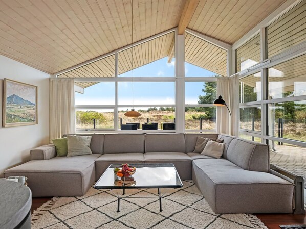 A modern living room with a sectional sofa, a glass coffee table, and large windows offering a view of the outdoors.