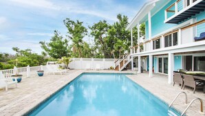 A swimming pool with a white deck, a covered patio area, and a view of the surrounding greenery. - Snook Away - Six bedroom bay front home nicely updated (Captiva)