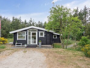 Een klein, donker houten huisje met een witte deur en ramen, nummer 23, omringd door bomen en een grindpad.