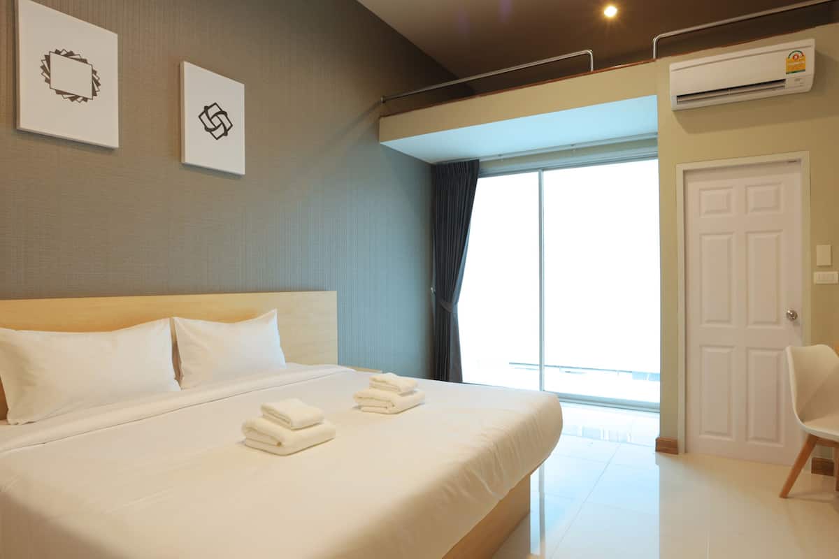 A hotel room with a large bed, a desk, and a chair. There is a window with curtains, an air conditioning unit, and a door.