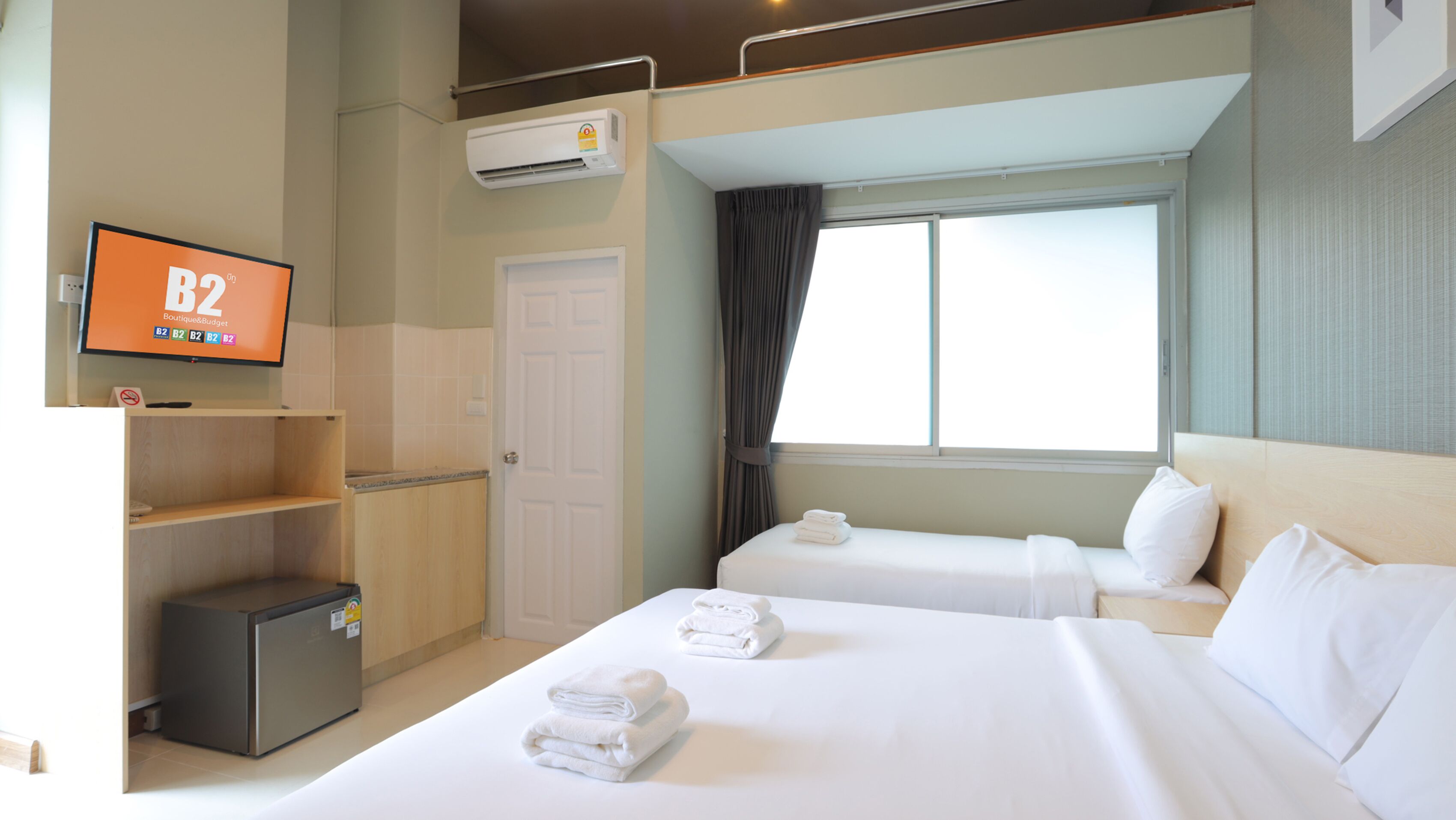 A hotel room with two beds, a TV on the wall, an air conditioning unit, and a window with curtains.