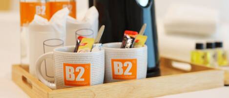 Una bandeja con dos tazas de la marca B2, una botella de líquido anaranjado, una cafetera y un recipiente pequeño con una cuchara.