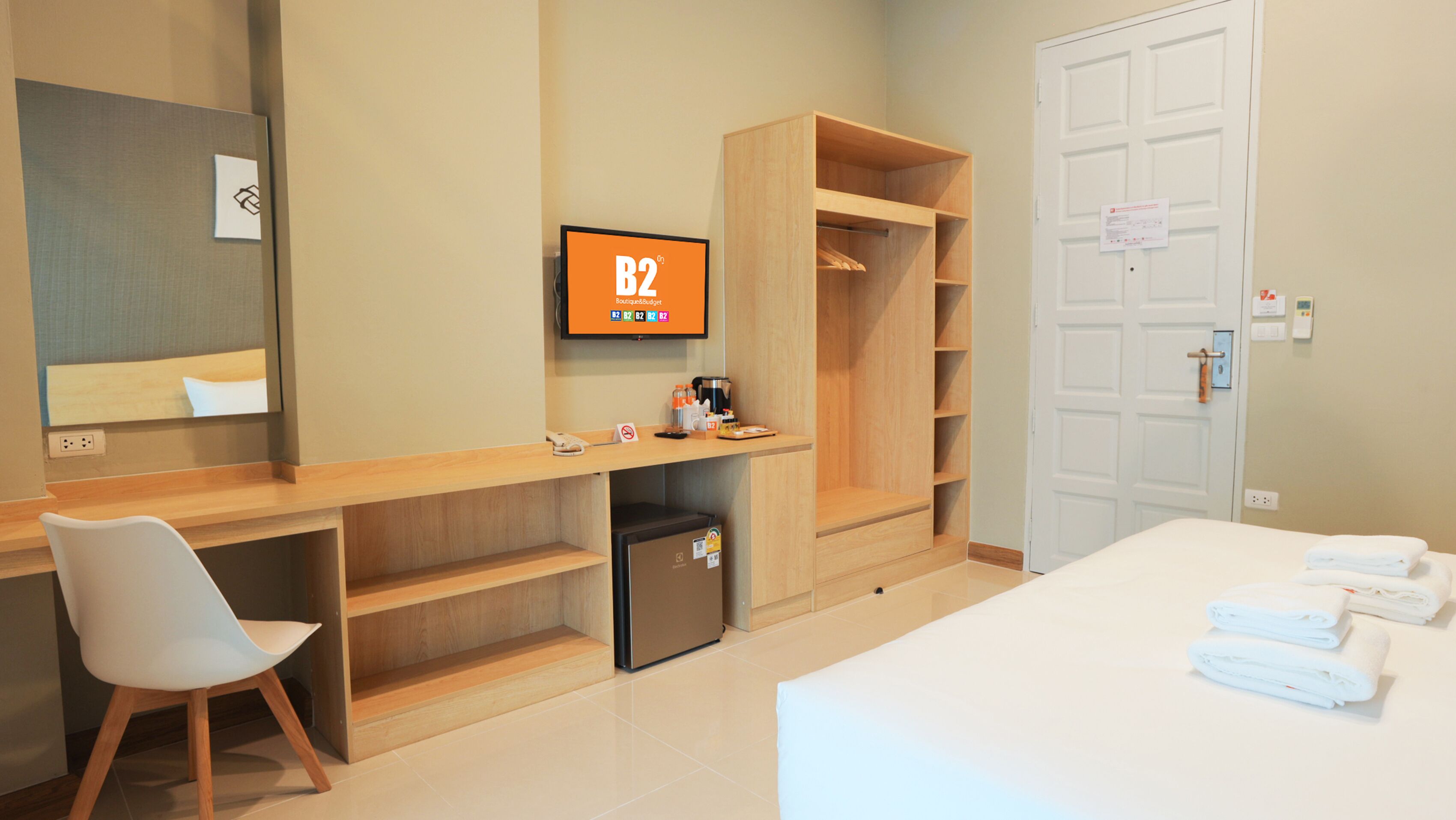 A hotel room with a bed, a desk, a chair, a wardrobe, a small refrigerator, and a television displaying 'B2 Boutique Budget'.
