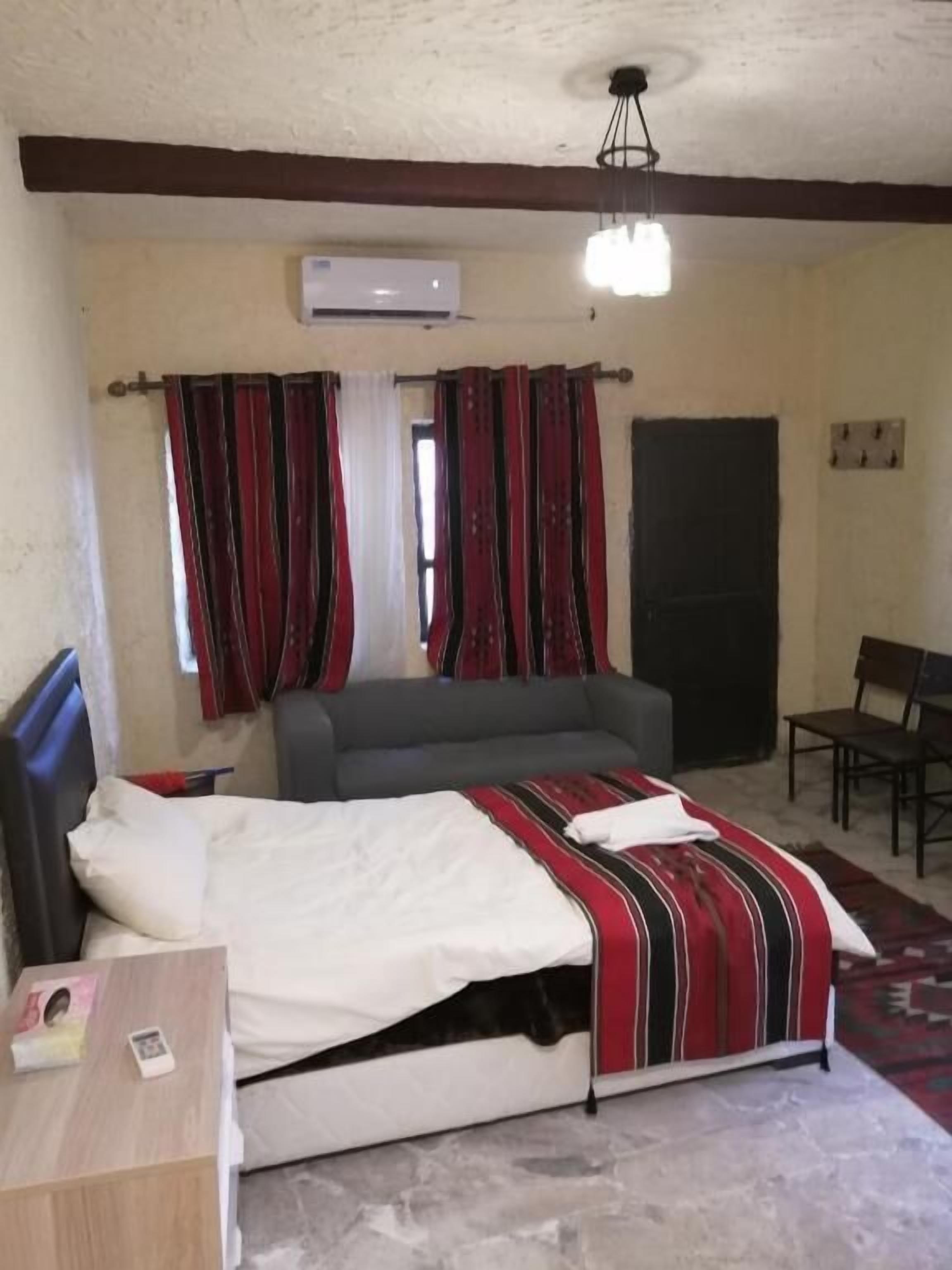 Ein Schlafzimmer mit Bett, einer Couch, einem Schreibtisch und einem Stuhl. Es gibt ein Fenster mit Vorhängen, eine Klimaanlage und eine Deckenleuchte.