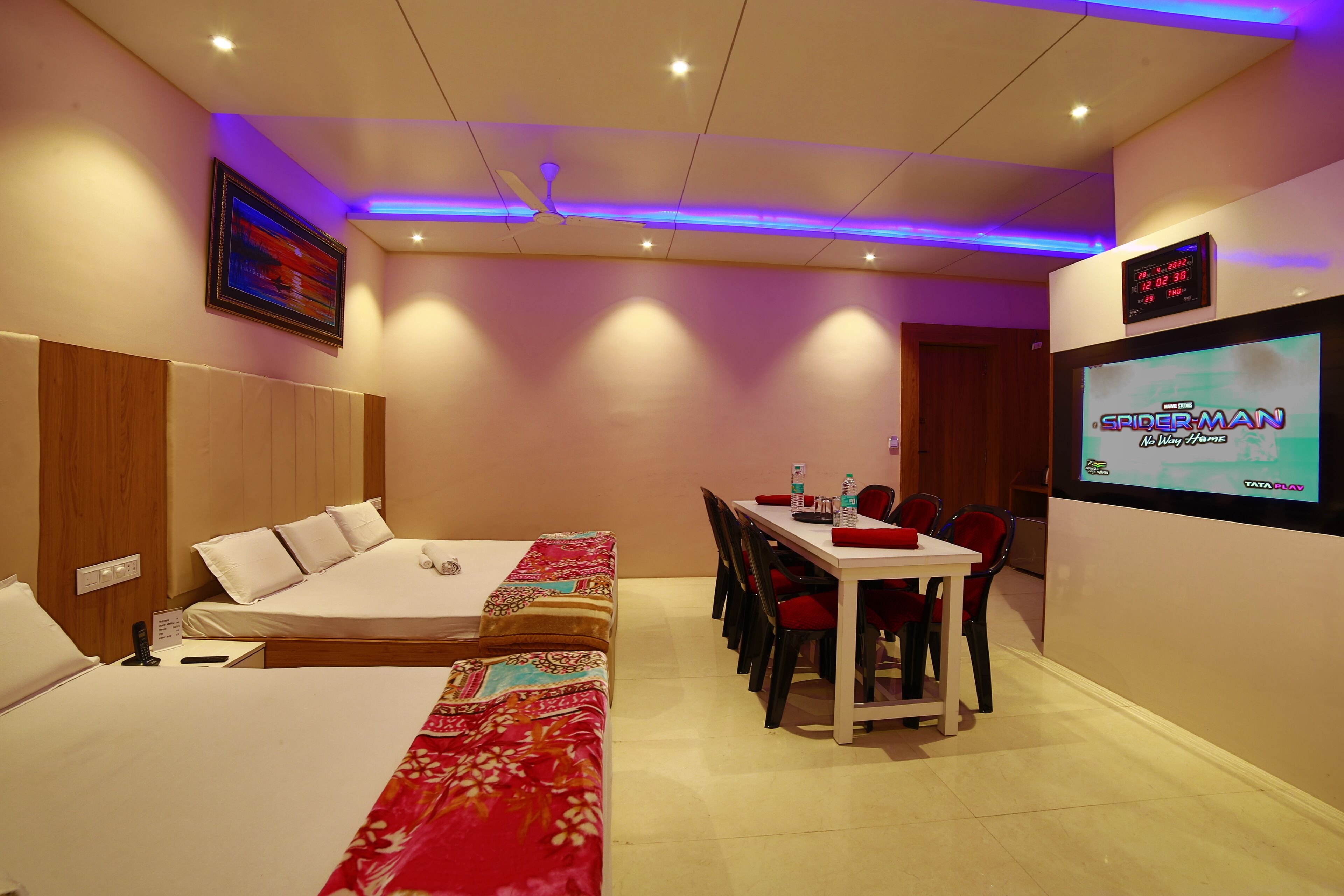 A hotel room with two beds, a dining table with chairs, a TV screen displaying 'Spider-Man: No Way Home', and a ceiling fan.