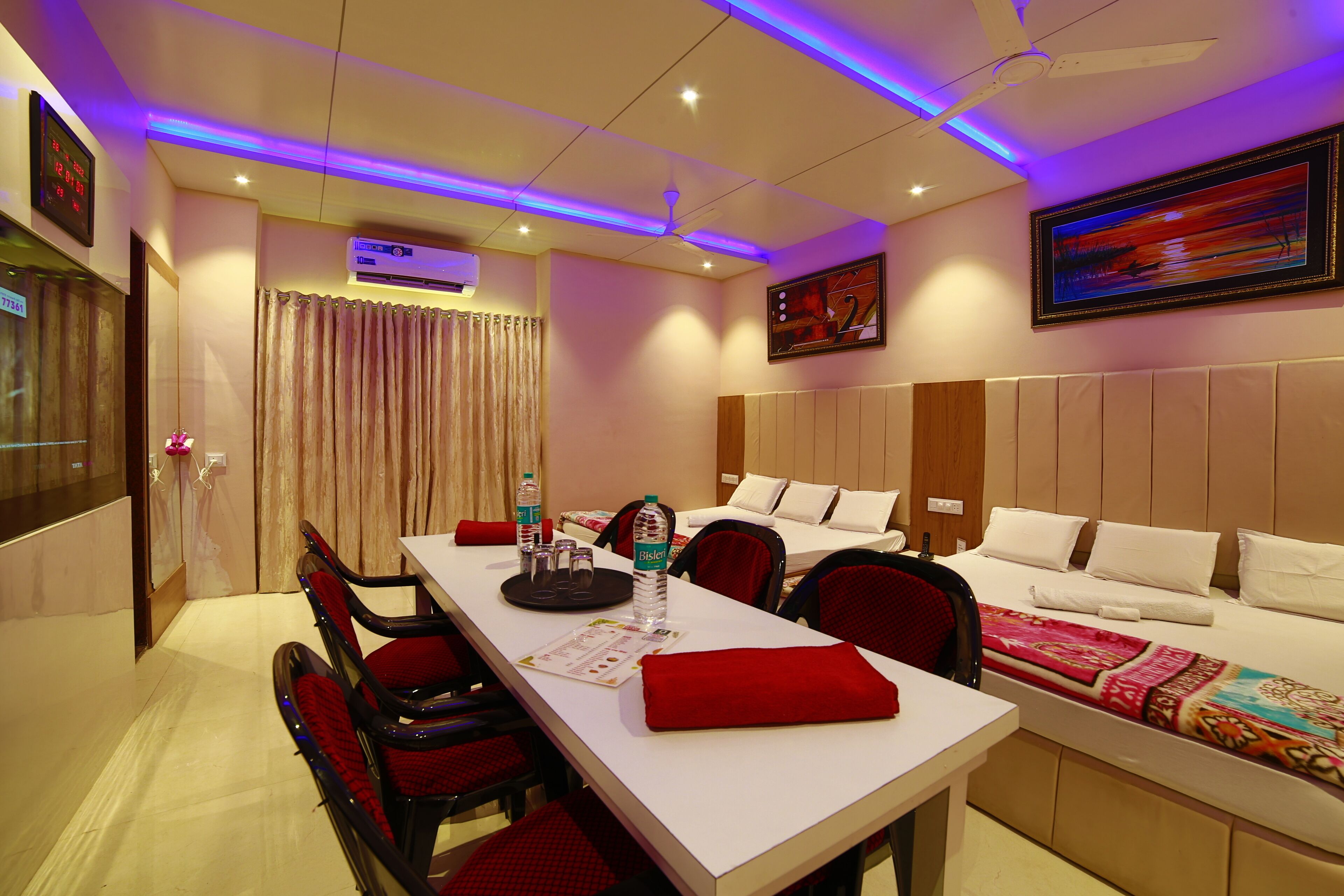 A hotel room with a bed, a desk with chairs, a table with a red cloth, and a painting on the wall.
