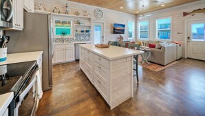 A spacious kitchen with white cabinetry, a central island, and a dining area with a gray sofa. - Pool Cabana, Backyard w/ Fire Pit, Walk to the Beach | Villa Reel by AvantStay (Port Aransas)