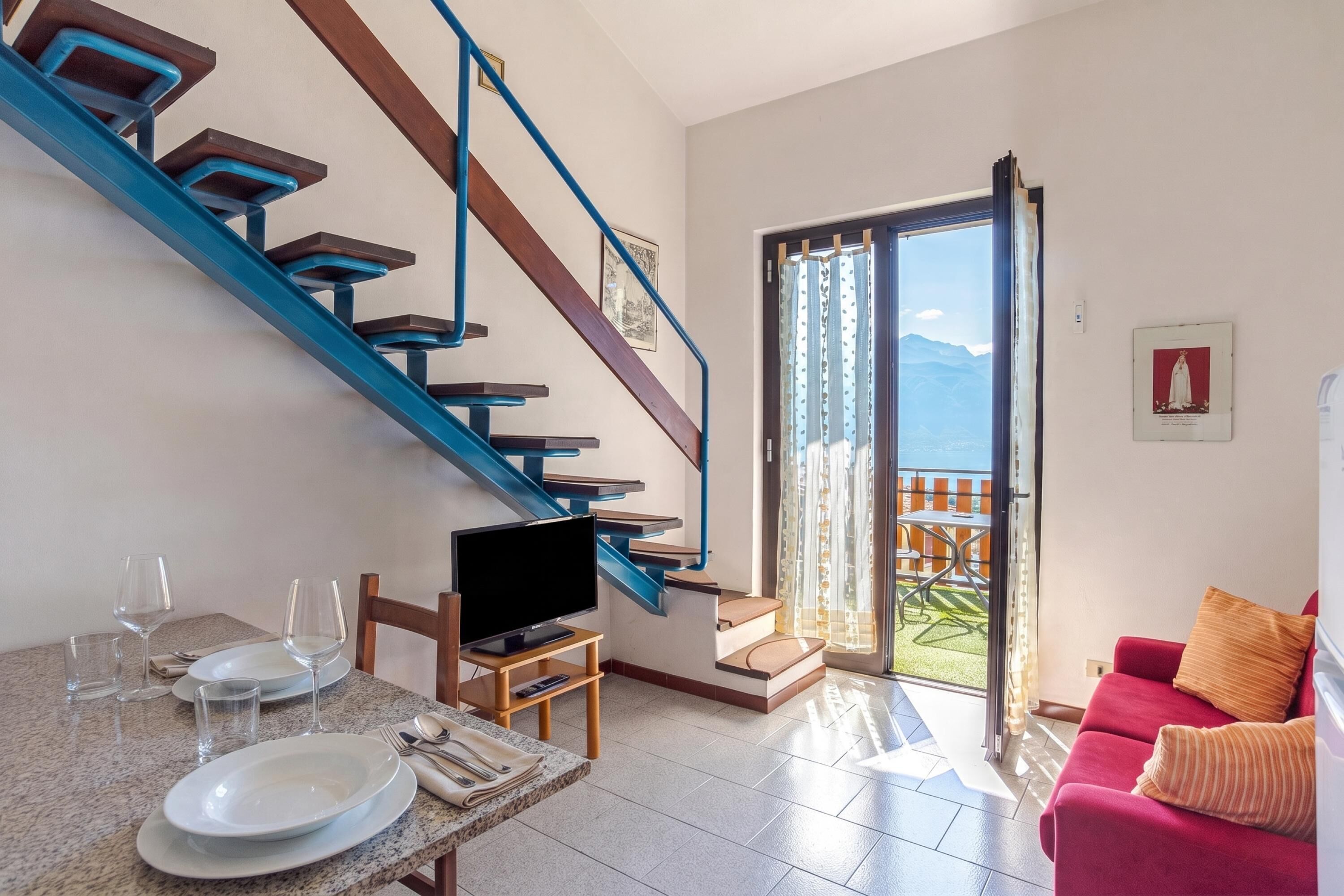 A modern living room with a blue staircase, a dining area with a table set for two, and a view of a balcony with outdoor furniture.