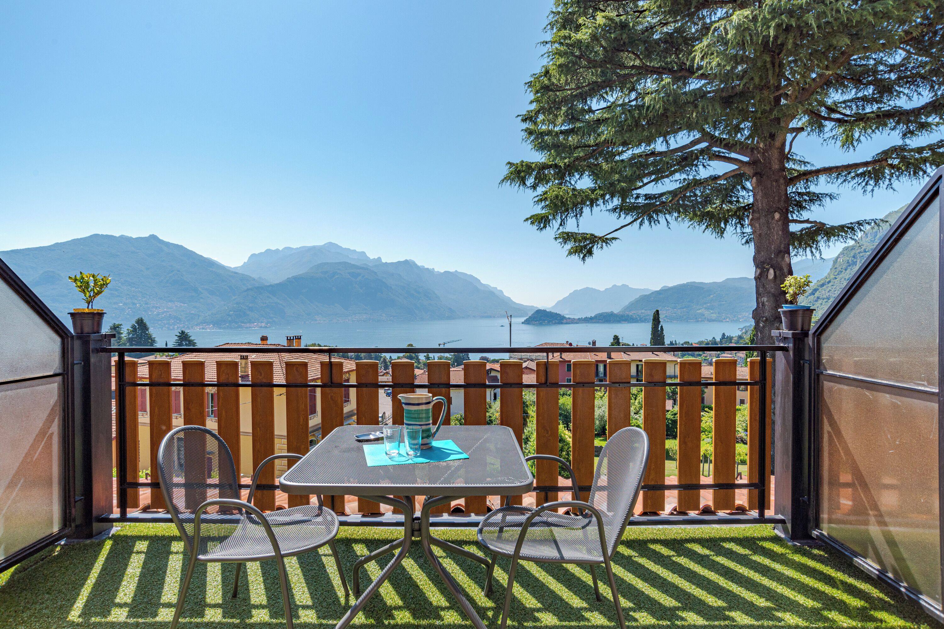 A balcony with a table and chairs, a wooden fence, and a view of mountains and a lake.