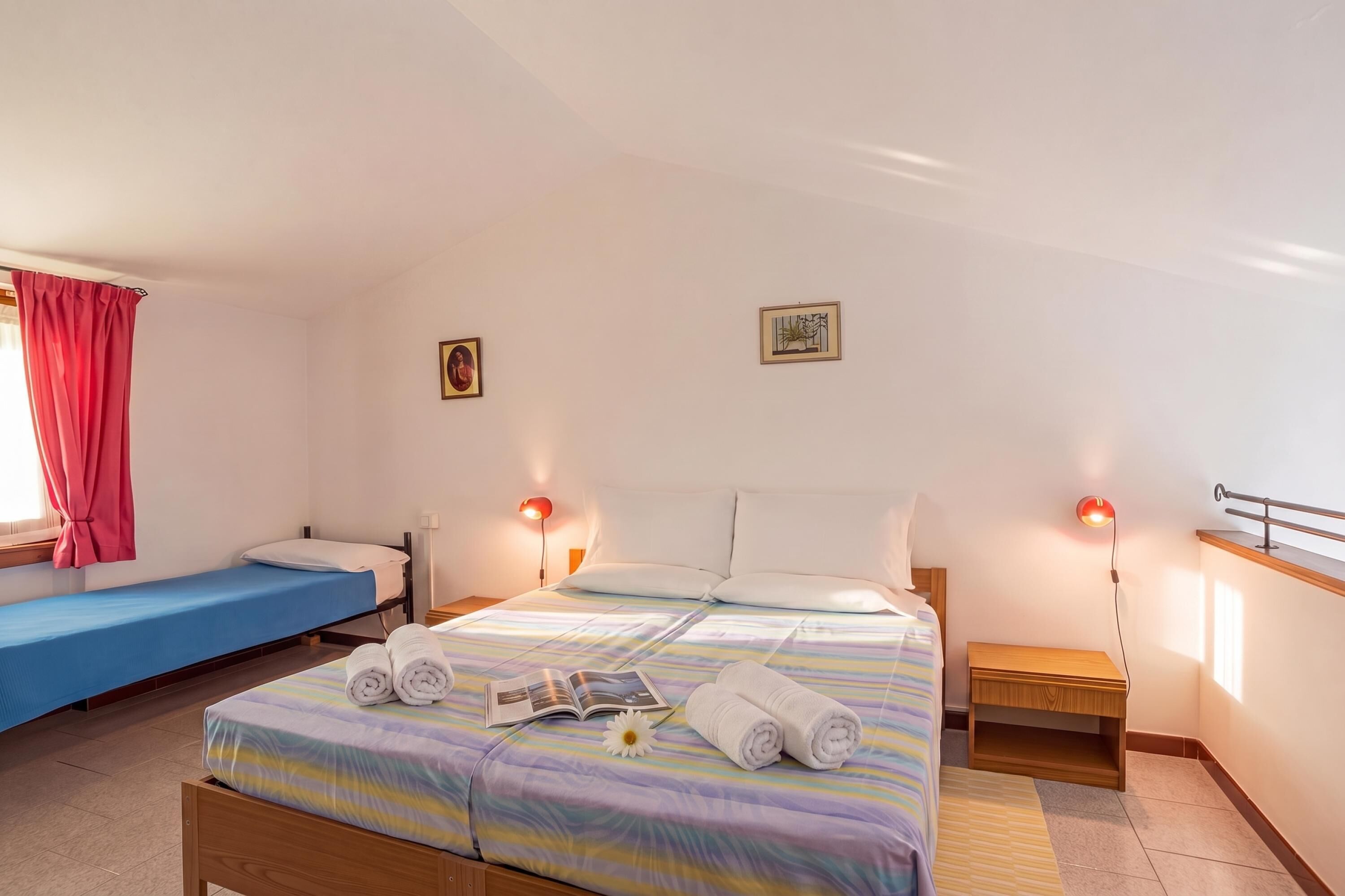 Une chambre d’hôtel avec un lit simple, un lit plus petit, une table de chevet avec une lampe, une fenêtre avec des rideaux et un tableau accroché au mur.