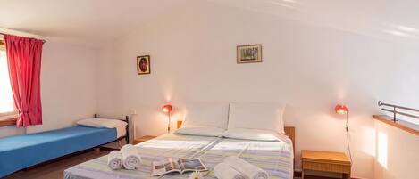 A hotel room with a single bed, a smaller bed, a bedside table with a lamp, a window with curtains, and a wall-mounted picture.