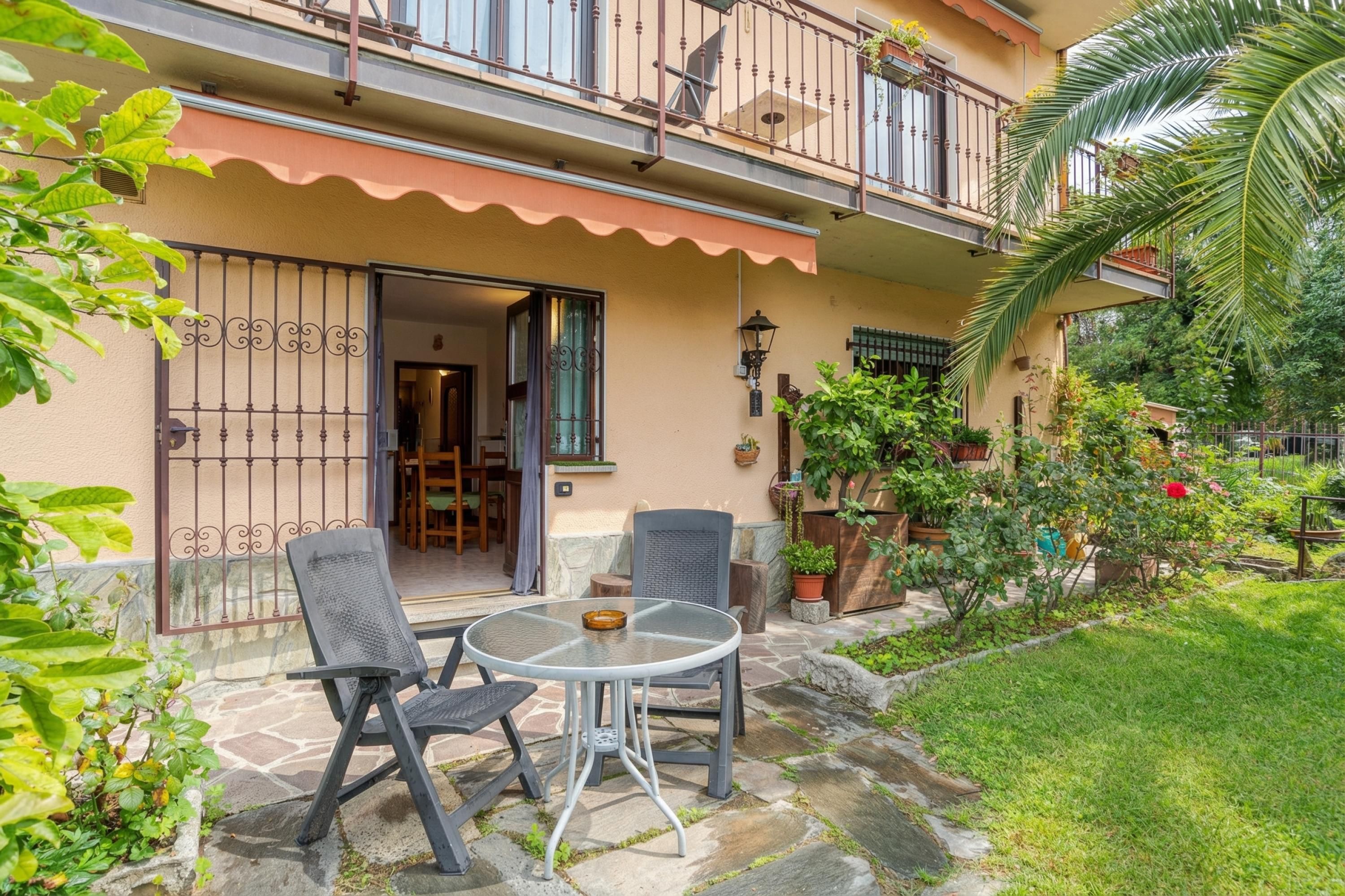 A two-story building with a balcony, a patio with a table and chairs, and a garden with potted plants.