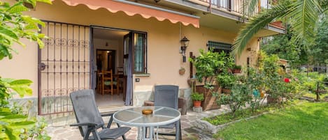 A two-story building with a balcony, a patio with a table and chairs, and a garden with potted plants.