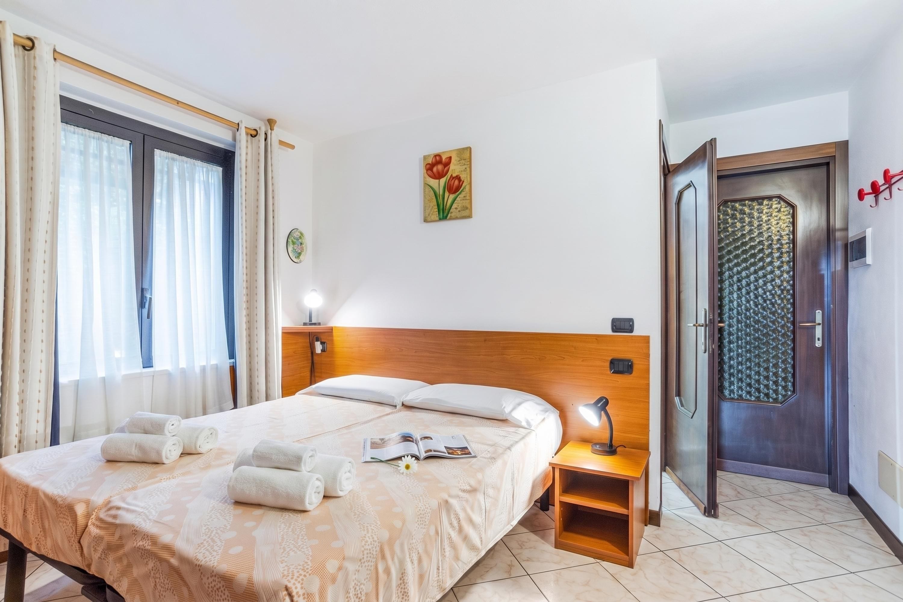 A hotel room with a bed, a bedside table with a lamp, a door with a textured glass panel, and a window with curtains.