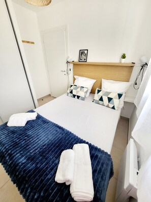 A neatly made bed with a blue blanket and two white towels, a wooden headboard, a small potted plant, and a framed picture on the wall. - L'échappée Belle en Tout Compris (Linguizzetta)