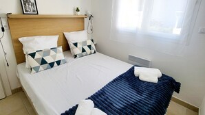 A neatly made bed with a blue blanket and two pillows, a bedside table with a lamp, and a window with curtains. - L'échappée Belle en Tout Compris (Linguizzetta)