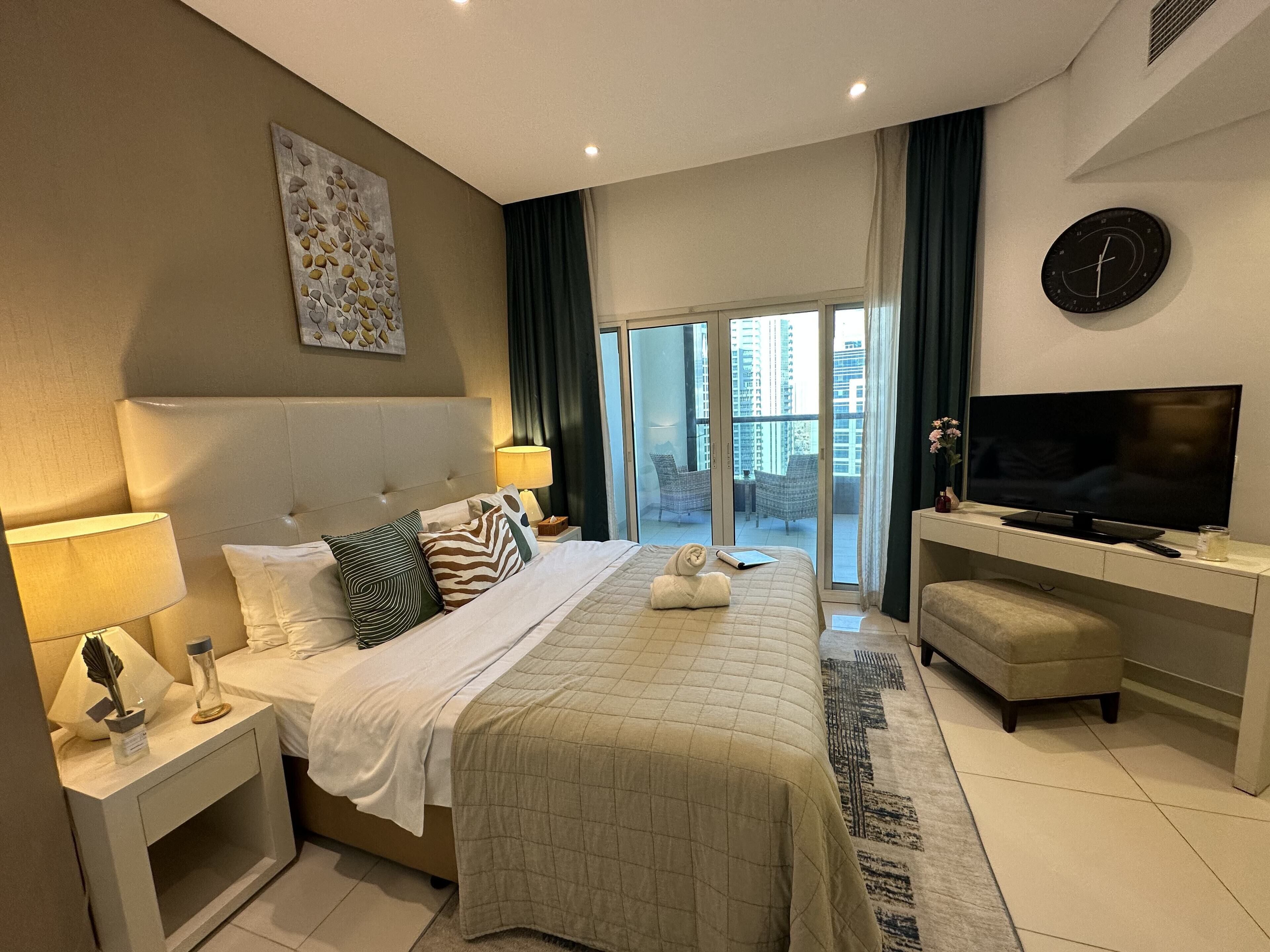 A modern hotel room with a large bed, a television, a desk, and a view of the city through the balcony.