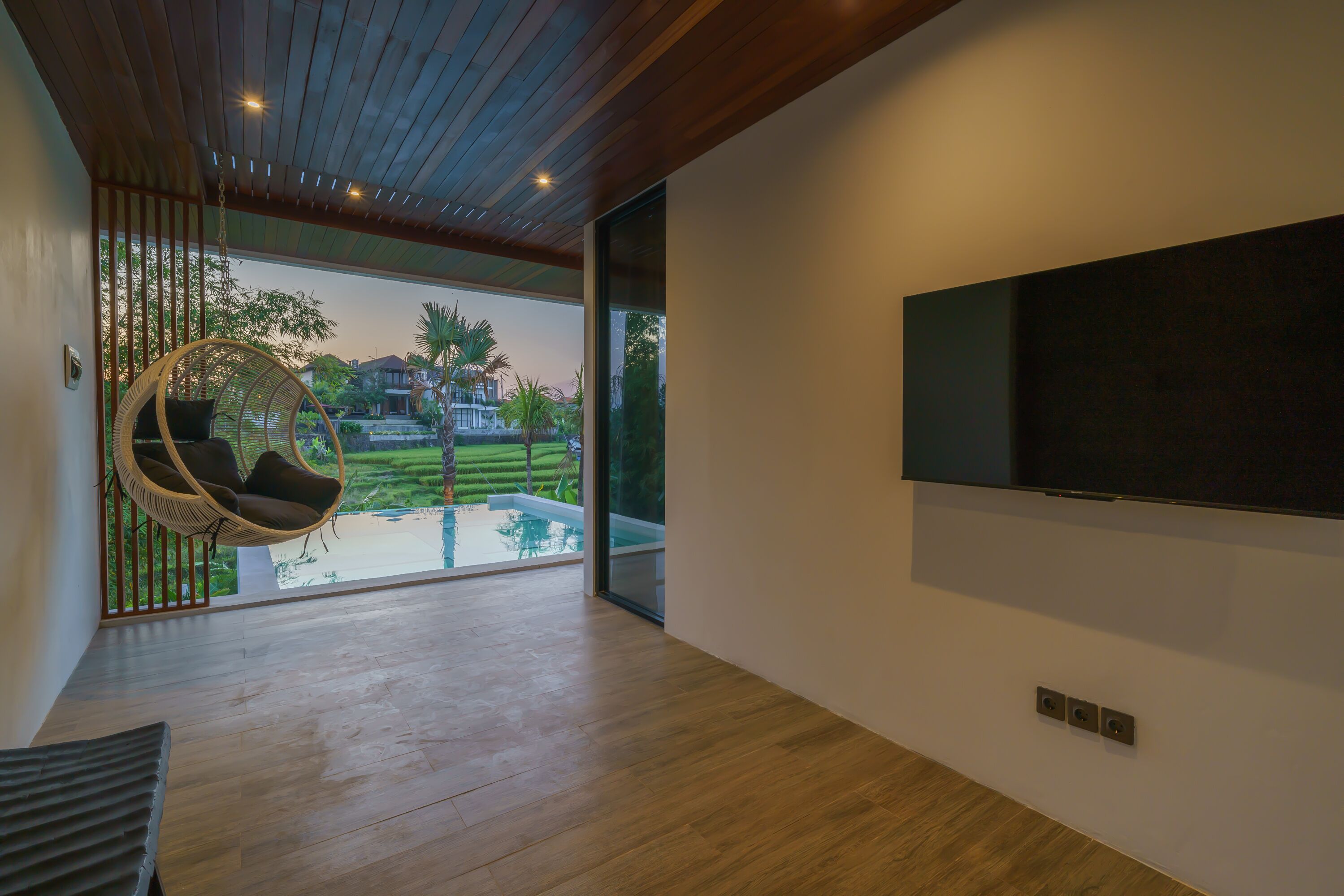 A modern interior with a wooden ceiling, a large TV on the wall, a hanging chair, and a view of a swimming pool and greenery outside.