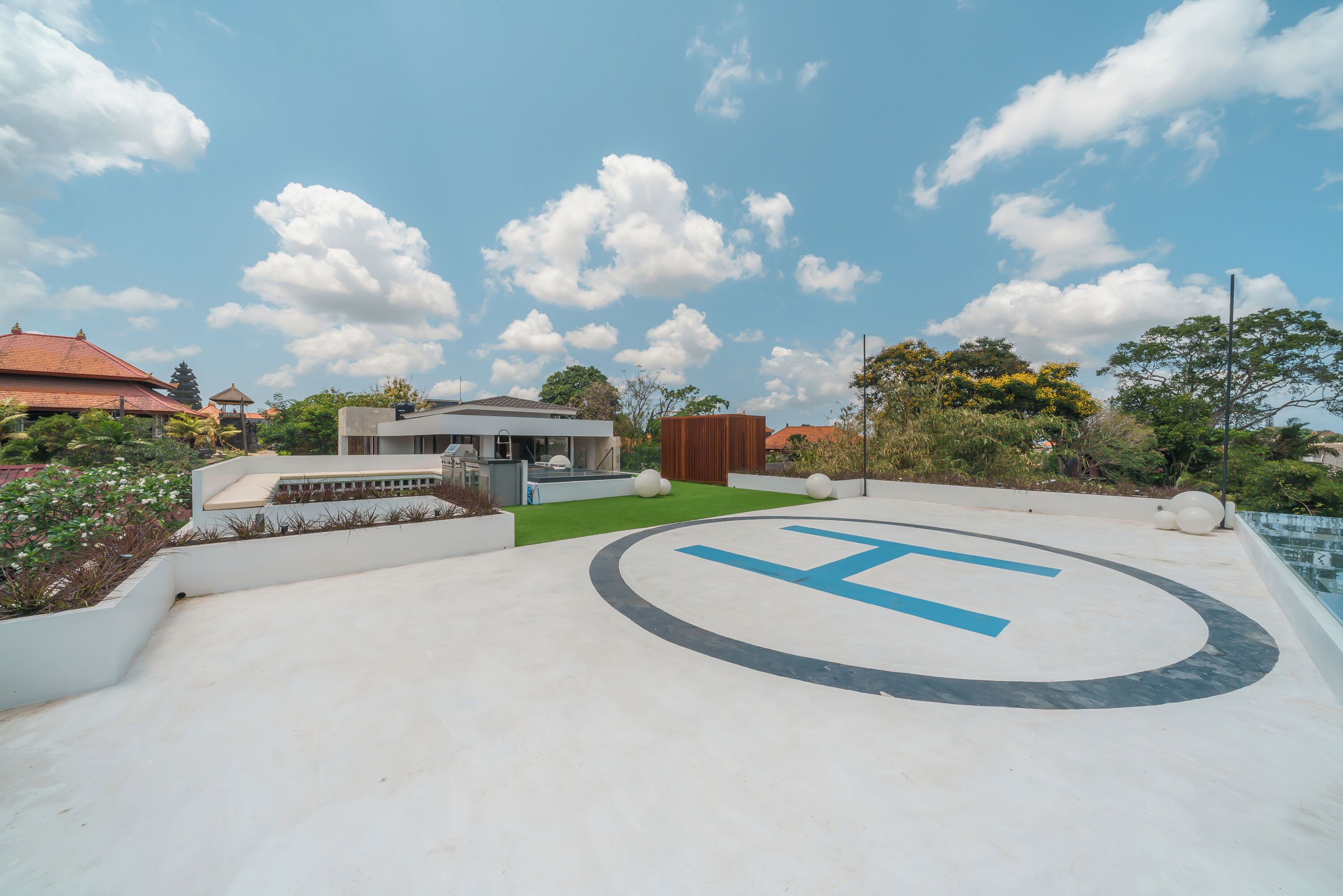 Un edificio moderno con un gran logo en forma de 'H' en el suelo, rodeado de vegetación y un cielo despejado.