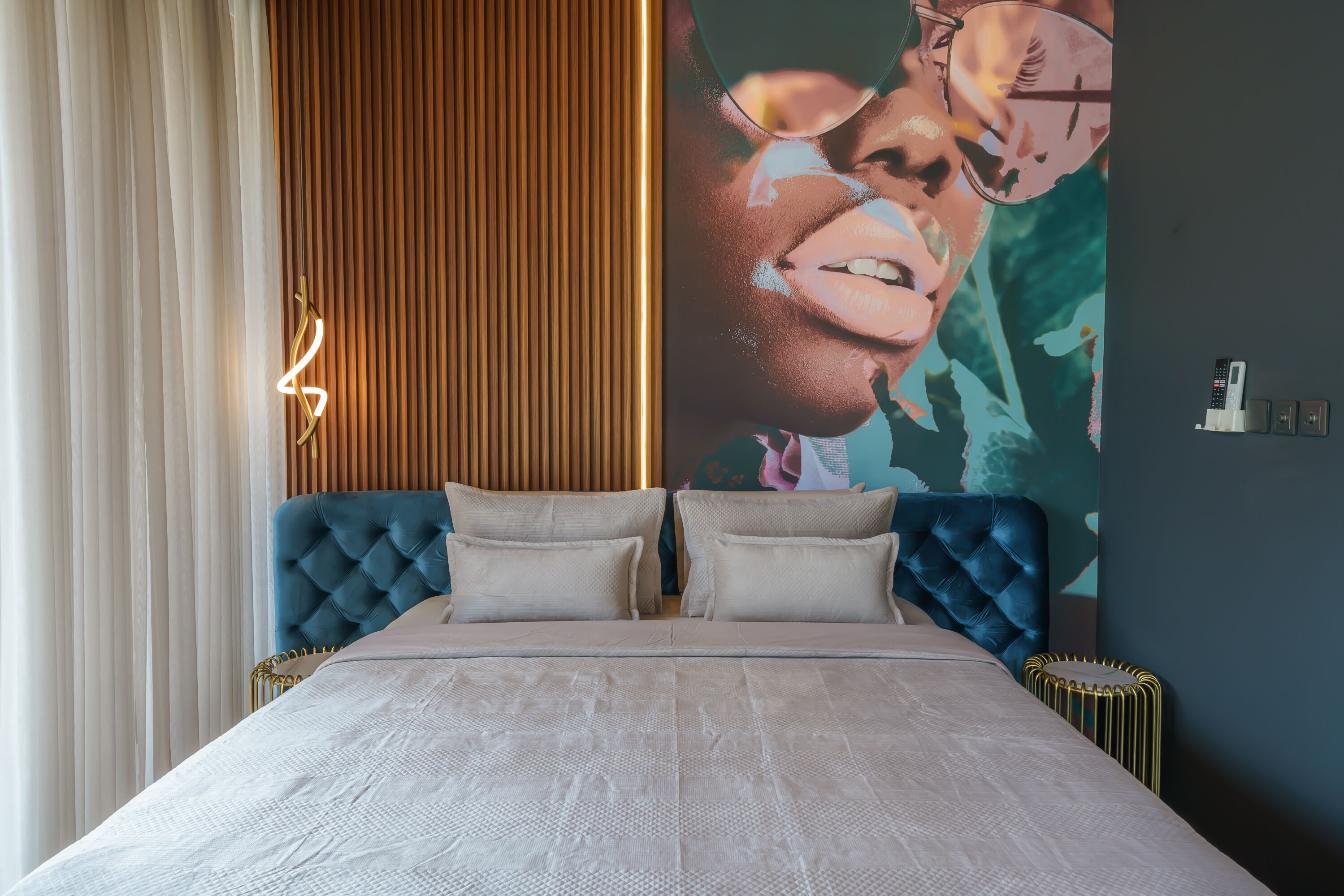 Une chambre à coucher avec une tête de lit capitonnée, un grand lit recouvert d’une couvre-lit à motifs et un luminaire fixé au mur.