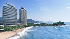 Un hotel costero con dos edificios destacados, una playa y vistas al mar y montañas a lo lejos.