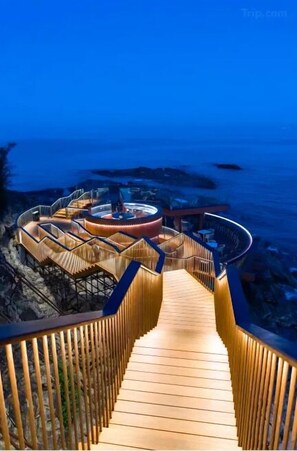 A wooden deck leading to a circular structure with a central seating area, overlooking a beach.