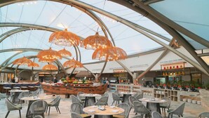 Un comedor interior con mesas y sillas redondas, una barra central de comida y un diseño arquitectónico moderno con techo de vidrio.