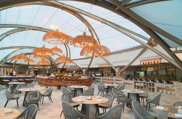 An indoor dining area with round tables and chairs, a central food display, and a modern architectural design with a glass ceiling.