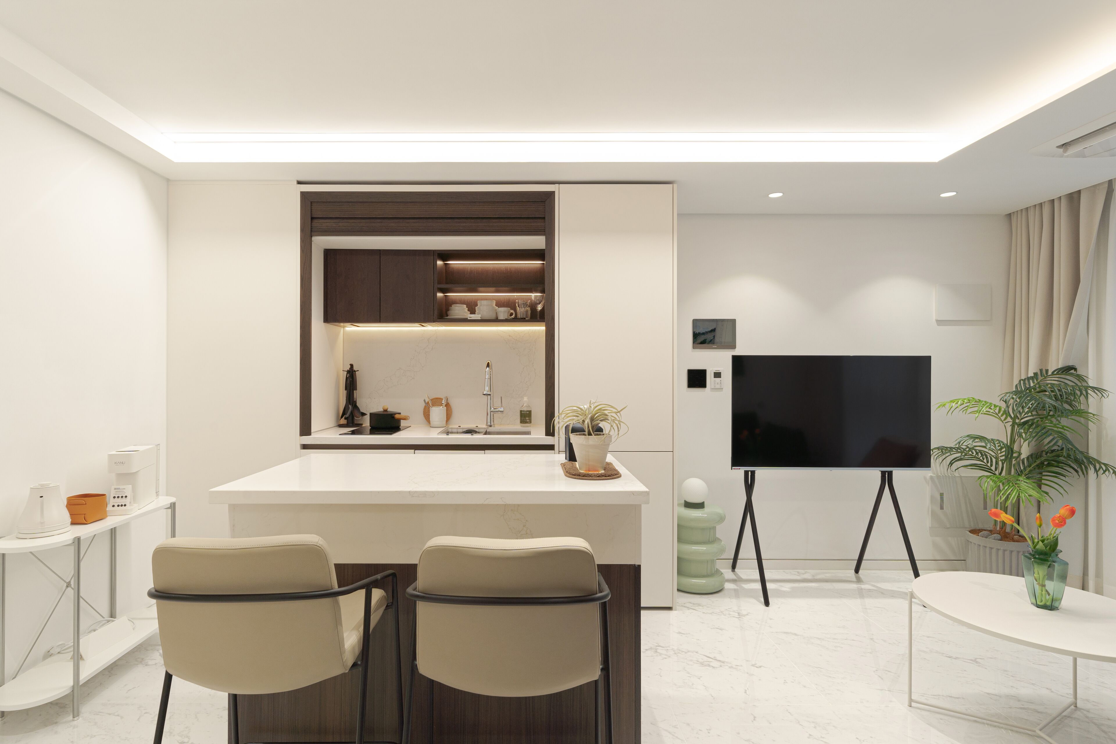Una cocina moderna con una isla central, dos taburetes de bar, una mesa de comedor y un televisor montado en la pared.