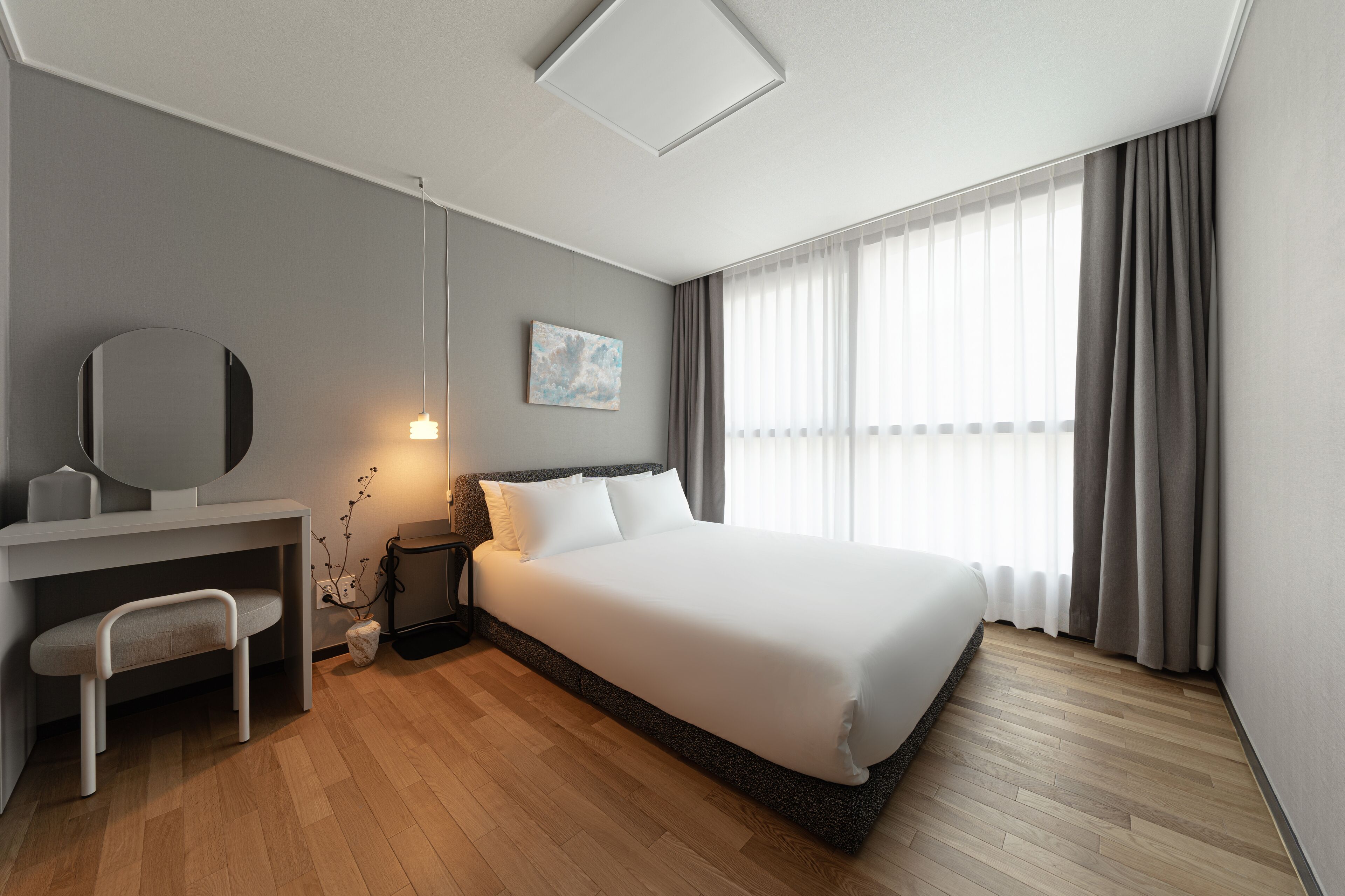 Una habitación de hotel moderna con una cama grande, un escritorio con silla, una mesita de noche y un ventanal con cortinas.