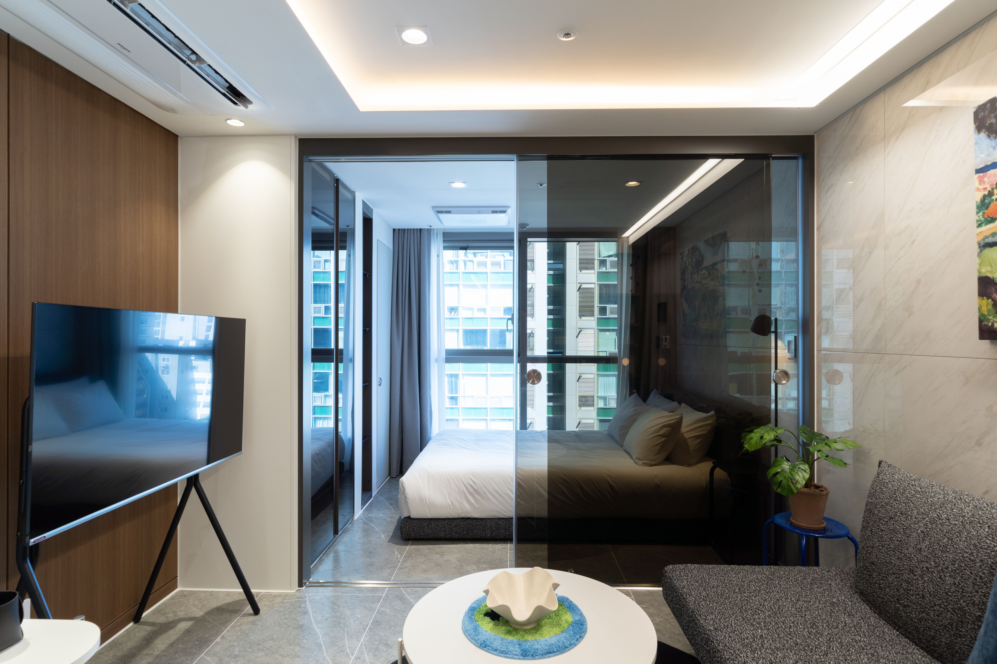 A modern hotel room with a large bed, a TV on a stand, a small round table, and a potted plant.
