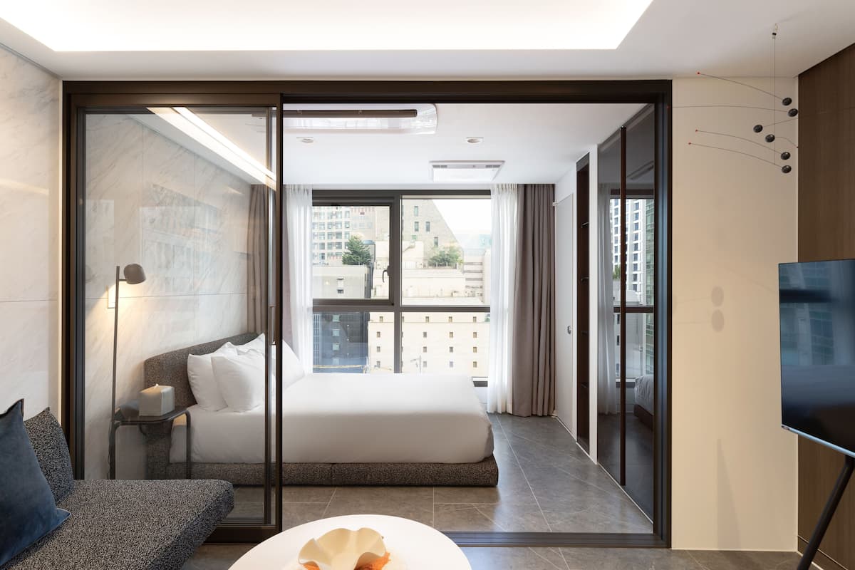A modern hotel room with a large bed, a sofa, a TV, and a view of the city through floor-to-ceiling windows.